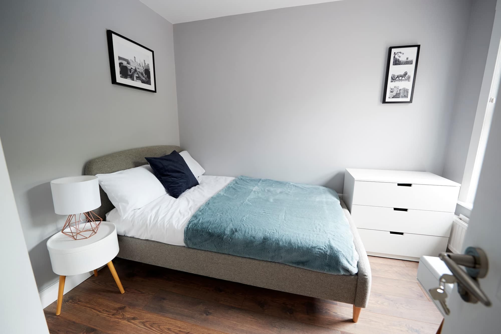 A small tidy bedroom with a grey fabric bed, white pillows, a teal throw and a white chest of drawers