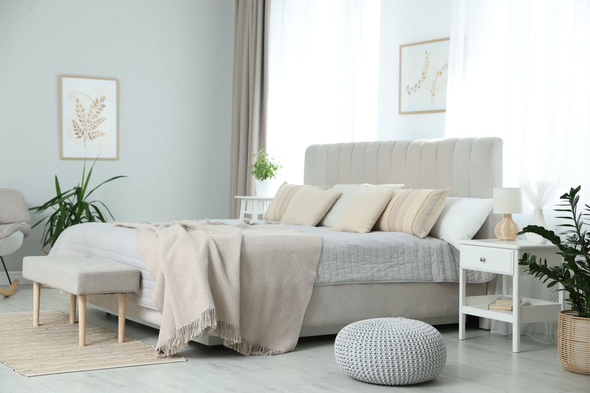 A bright, calm bedroom with a beige upholstered bed, layered cushions, a knitted throw and soft window light