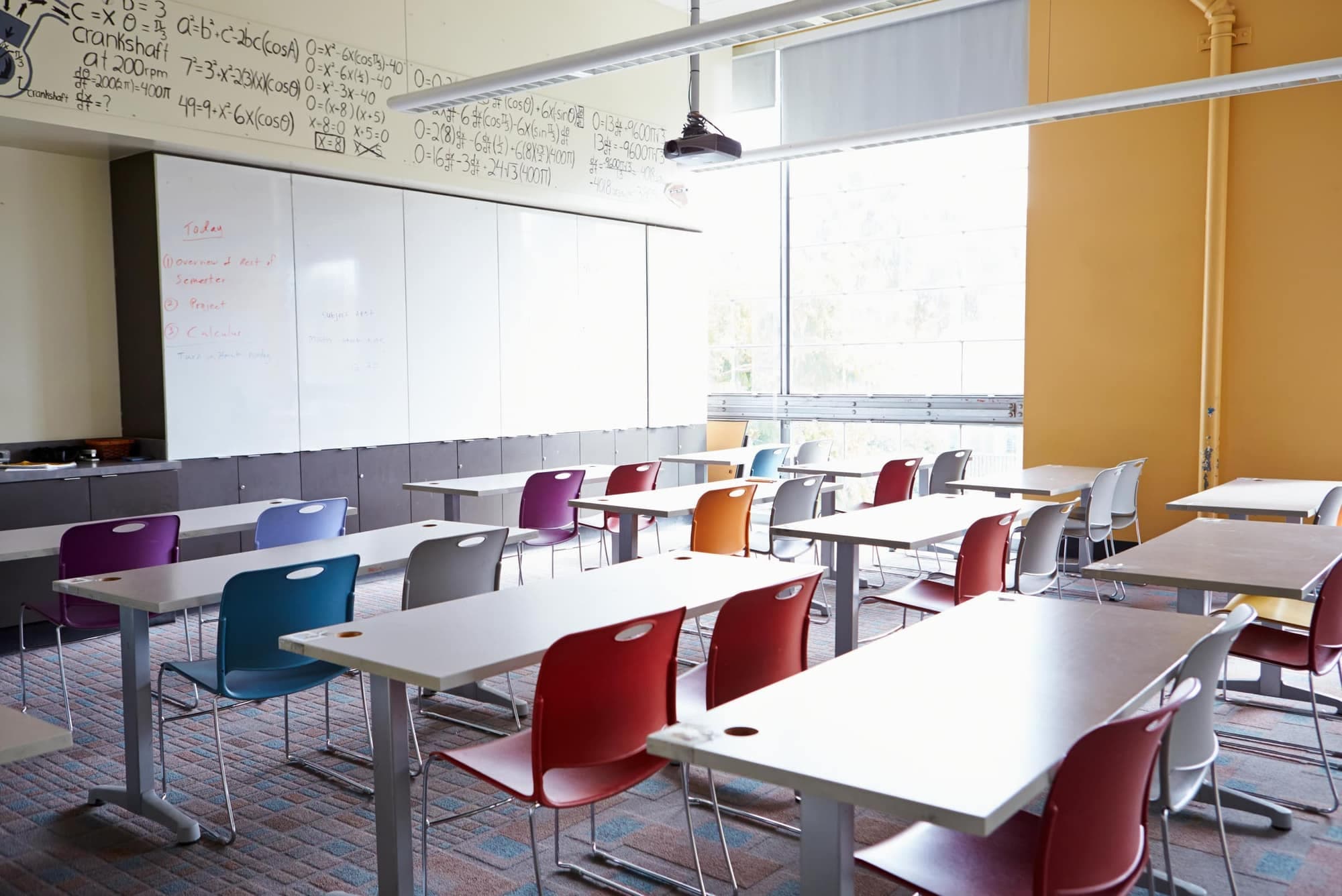 An empty classroom with white desks, colourful chairs, a whiteboard and a large sunny window