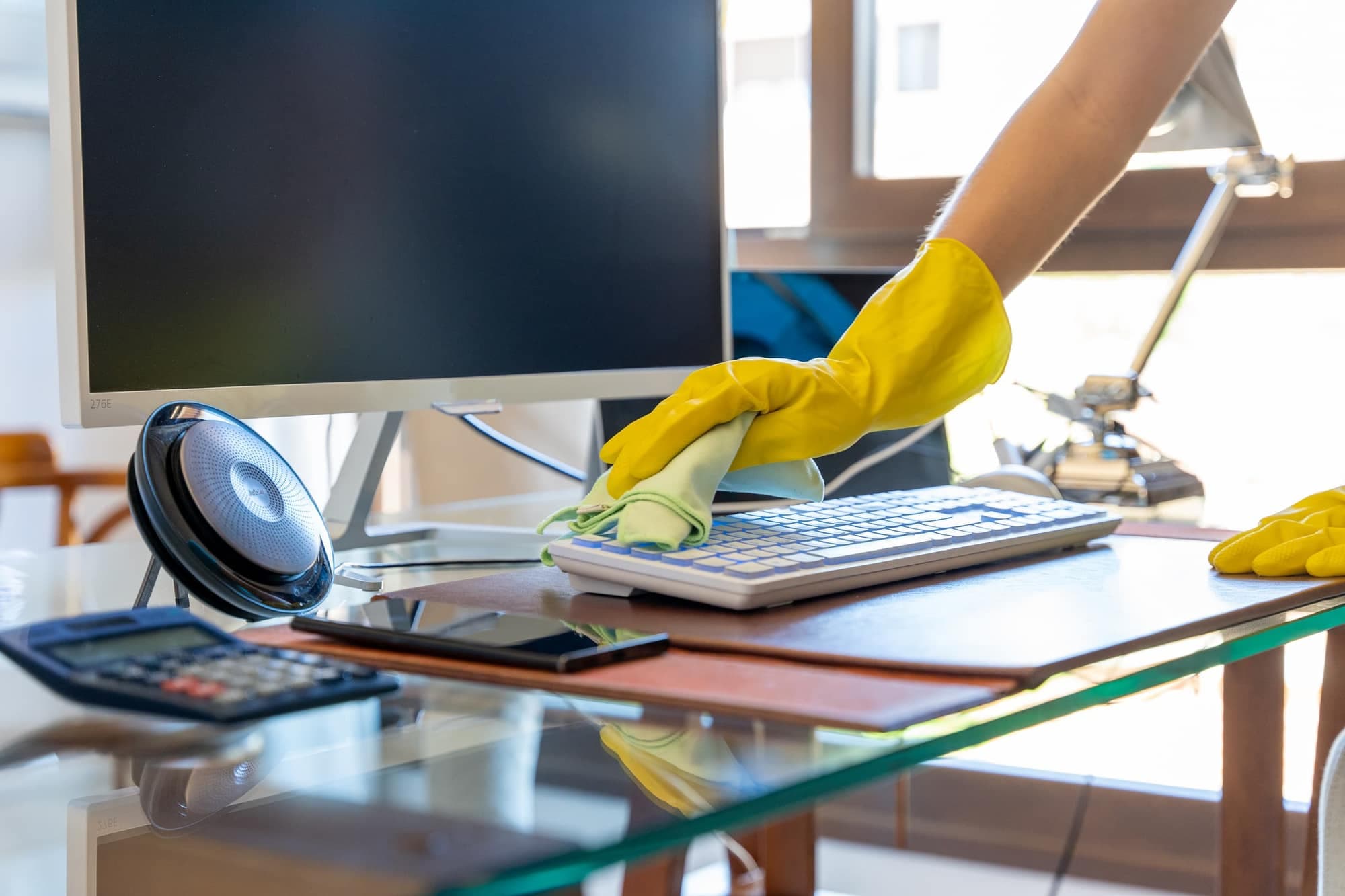 A hand in a yellow rubber glove wipes a keyboard with a green cloth on a glass office desk
