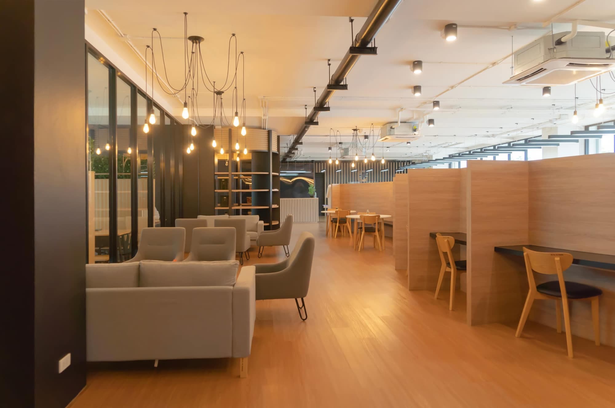 A warmly lit open-plan coworking space with grey sofas, hanging bulb lights, wooden booths and a polished wood floor
