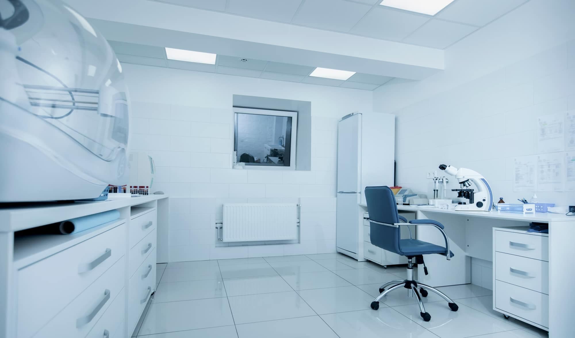 A spotless white laboratory with tiled walls, a microscope on a bench, a blue office chair and a shiny tiled floor