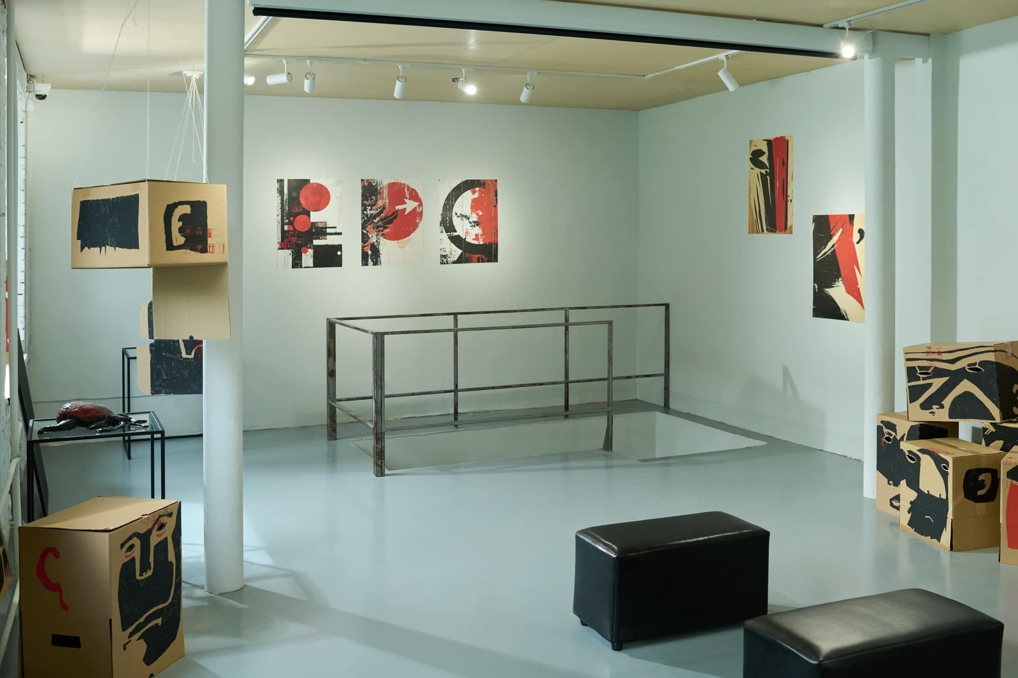 A pale gallery room with abstract posters on the wall, a metal railing around a floor opening and black leather ottomans