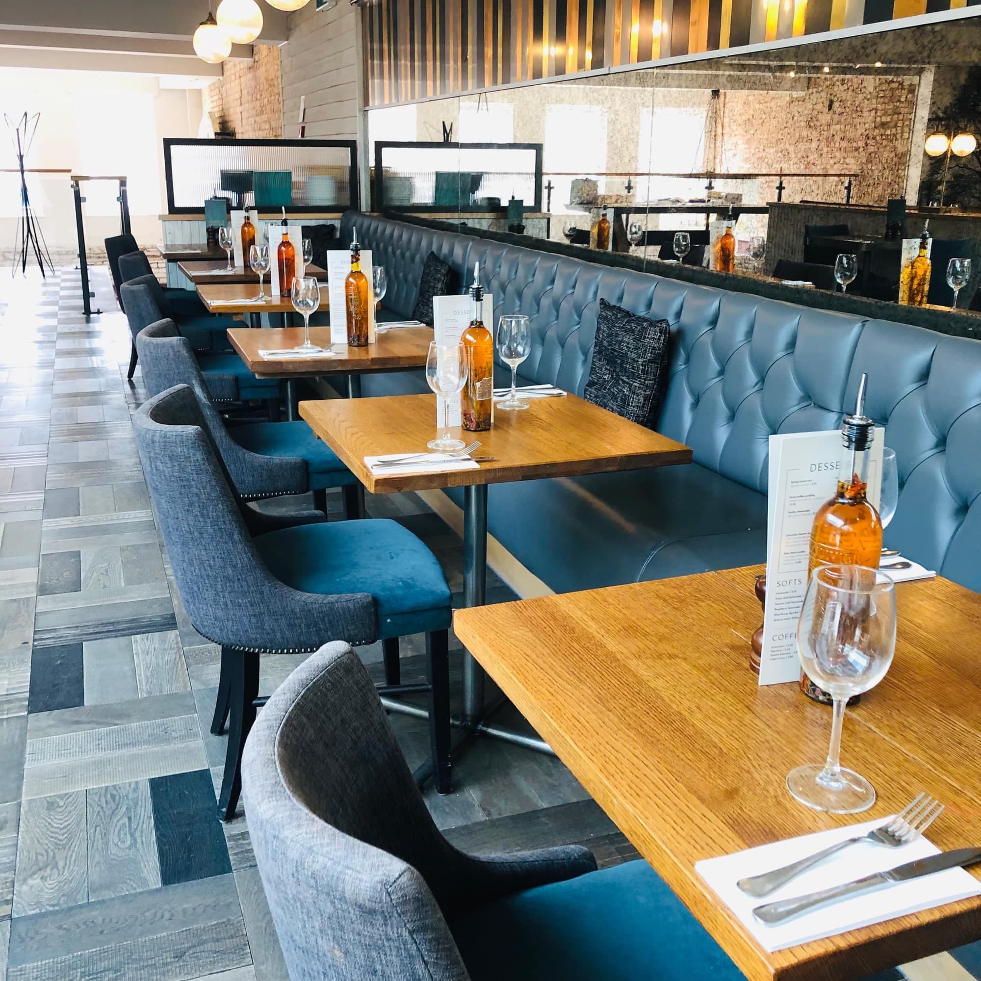Wooden restaurant tables set with wine glasses and menus beside a blue tufted leather banquette